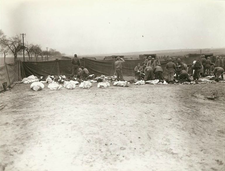 Soldados estadounidenses y alemanes muertos en un cementerio antes del entierro, lugar desconocido. Cada cuerpo fue colocado en una funda de colchón. Varios prisioneros alemanes cavan las tumbas y colocan cadáveres en ellas