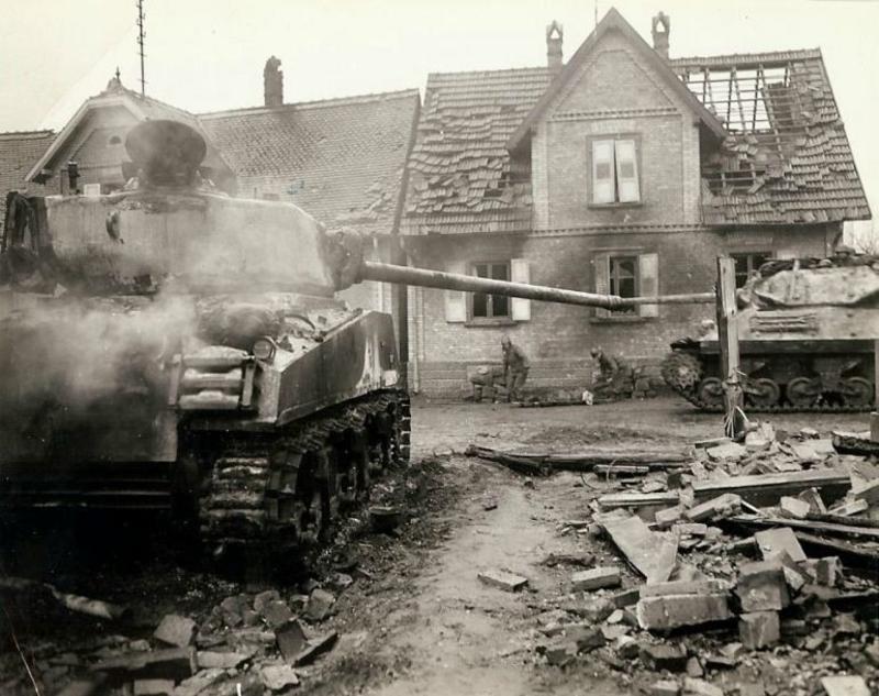 Un destructor de tanques junto con un tanque estadounidense destrozado durante la batalla por una ciudad. Entre los dos vehículos blindados se ven dos enfermeros que recogen el cuerpo de un soldado estadounidense que murió en la lucha por la urbe